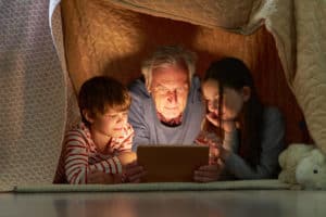 Children and grandfather using tablet computer or e-book under a blanket as a tent
